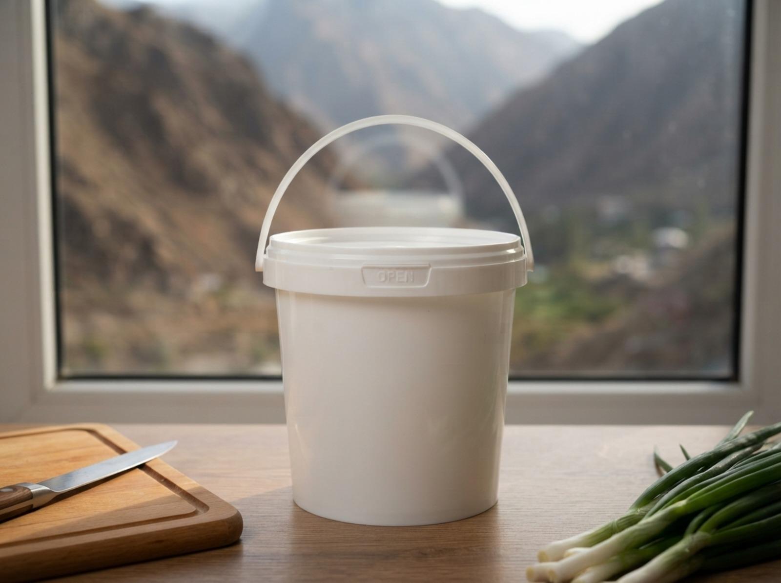 White 900 ml plastic bucket with handle and lid on a table by a kitchen window in Kara-Kul, with the mountains of the Naryn Valley outside