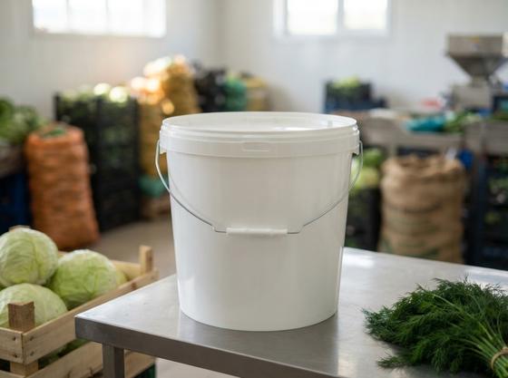 White 21 L plastic bucket with lid and metal bail handle on a steel table in a vegetable processing room in the Chuy Valley, next to a crate of cabbage