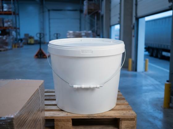 White 11.5 L plastic bucket with metal bail handle and lid on a wooden pallet in the shipping area of a factory warehouse by the open gates