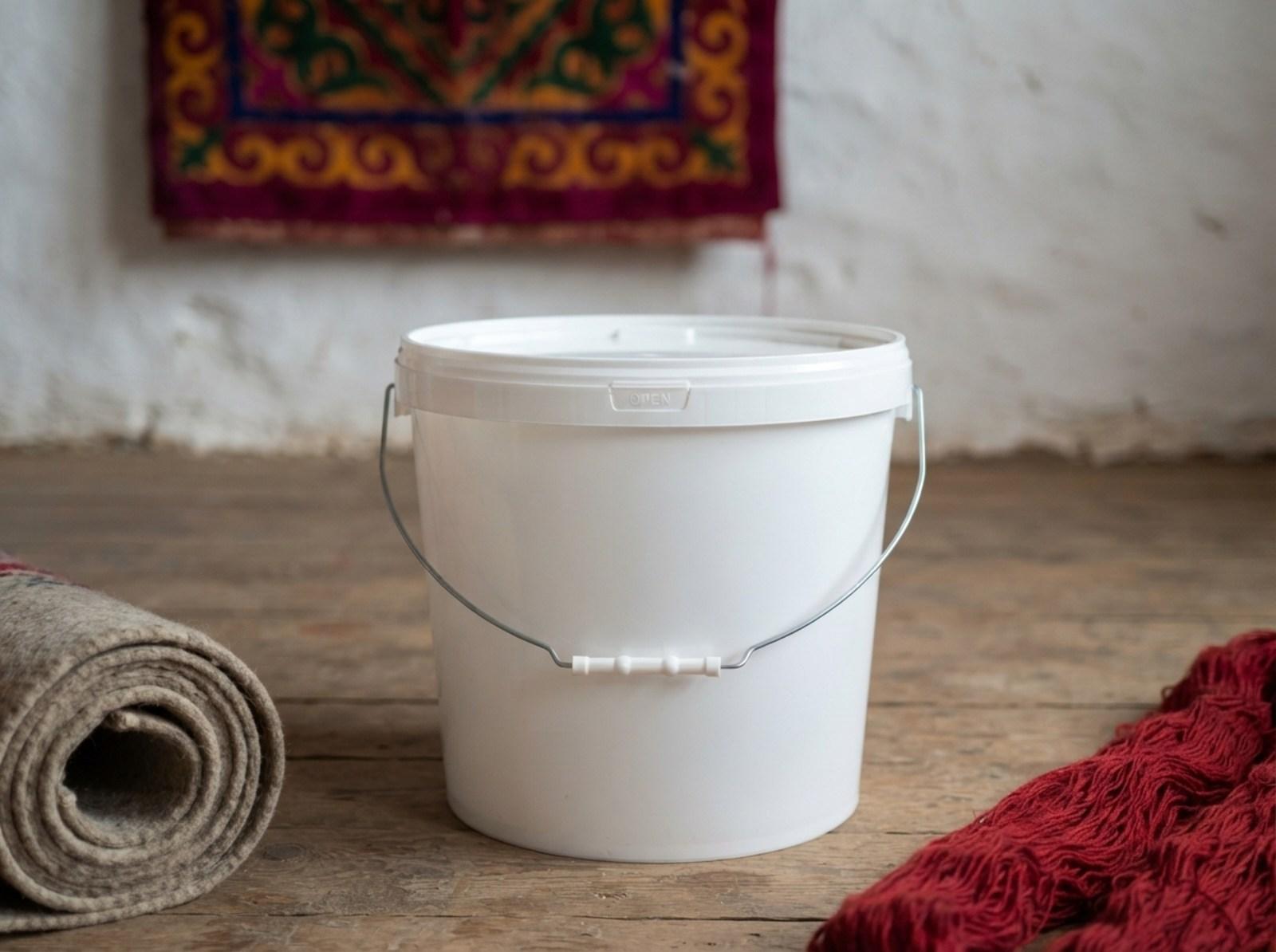 White 15 L plastic bucket with lid and metal bail handle with plastic grip on the wooden floor of a felt workshop in Kochkor, next to a roll of felt and dyed wool, with a shyrdak on the wall