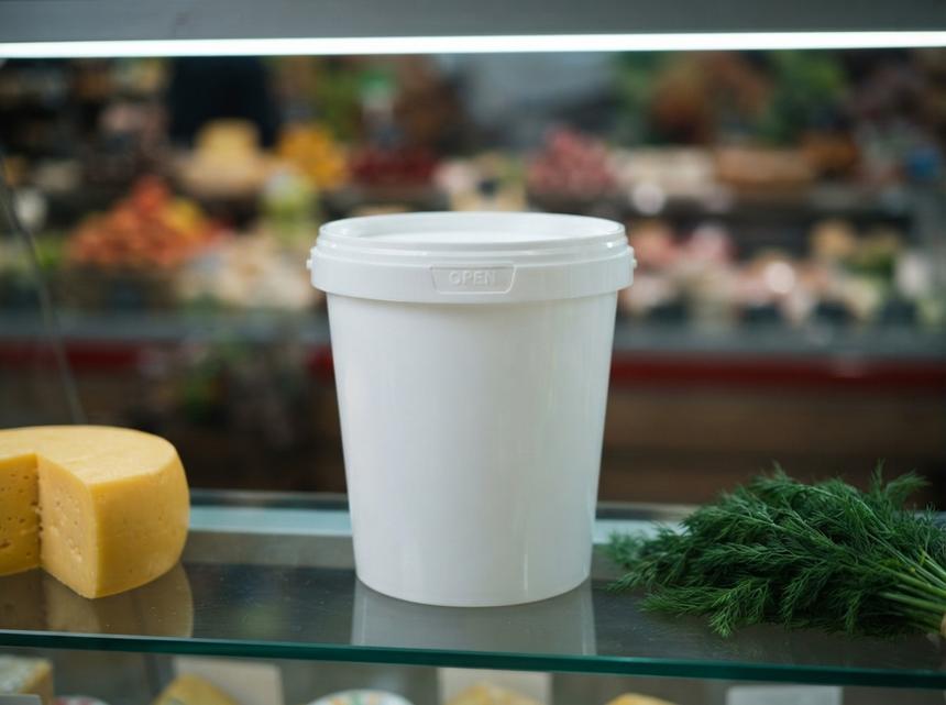 White 850 ml plastic bucket with lid on a glass shelf in a refrigerated store display case in Almaty, next to a wheel of cheese