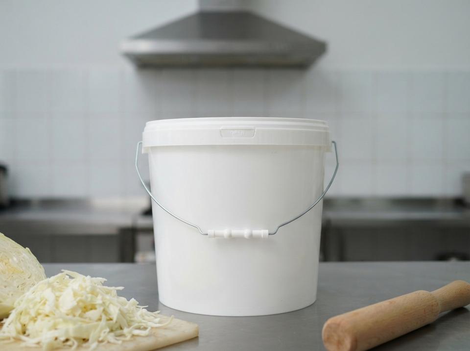 White 15 L plastic bucket with lid and metal bail handle with plastic grip on a steel table in a canteen kitchen, next to a pile of shredded cabbage