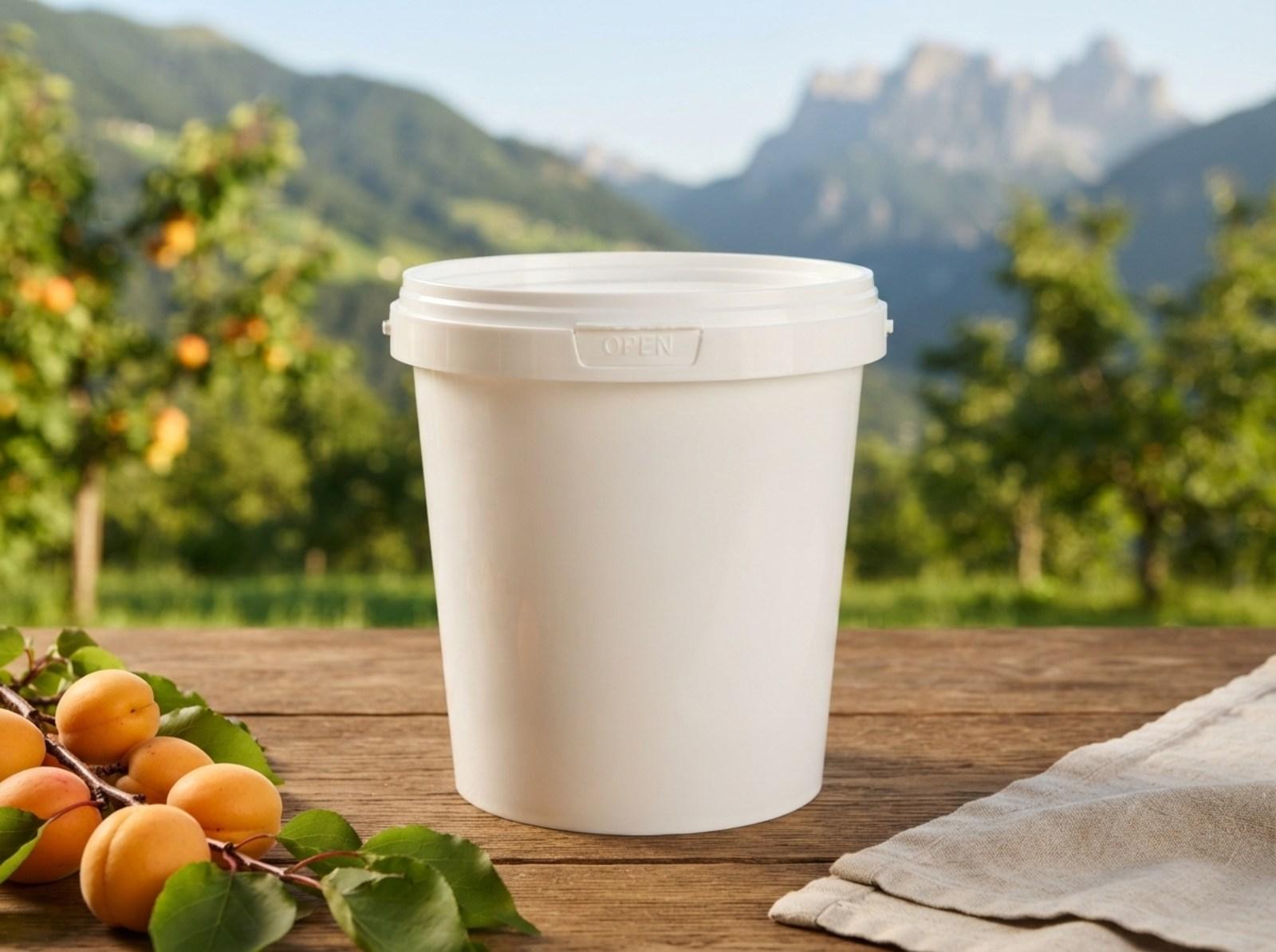 White 850 ml plastic bucket with lid on a garden table on a terrace next to a branch of ripe apricots, with mountain slopes covered in fruit trees behind