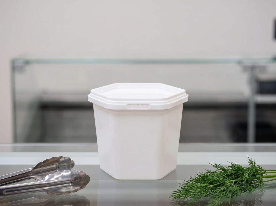 阿斯塔纳熟食店玻璃展柜中的 400 毫升白色六角形带盖塑料食品盒，旁边是食品夹和一枝莳萝
